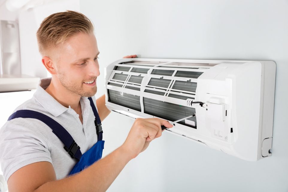 A smiling technician in blue dungarees servicing the front panel of a white wall-mounted split air conditioning unit