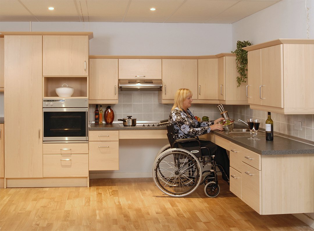 A woman in a wheelchair working at a light-wood kitchen with a lowered worktop and clear knee space beneath it